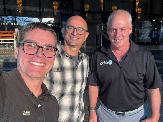three men standing shoulder to shoulder. The left person wears dark-rimmed glasses and a charcoal polo; the middle person wears glasses and a gray plaid button-down; the right person wears a black polo with the Aira logo.