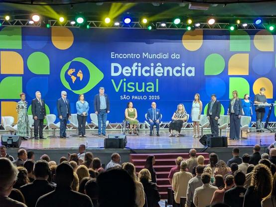 A straight-on photo of a group of presenters standing and seated across the stage. One presenter uses a wheelchair near center. The backdrop reads “Encontro Mundial da Deficiência Visual — São Paulo 2025,” with colorful geometric shapes. Stage lights in multiple colors frame the scene, and the audience is visible from behind.