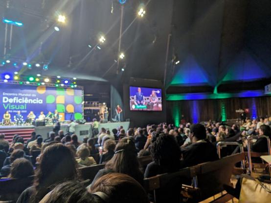 From the back of a large hall, rows of wooden-framed seats are filled with attendees. The stage is lit with green, blue, and yellow lights. Big screens on both sides show panelists. A large backdrop reads “Encontro Mundial da Deficiência Visual,” with abstract green and yellow shapes.
