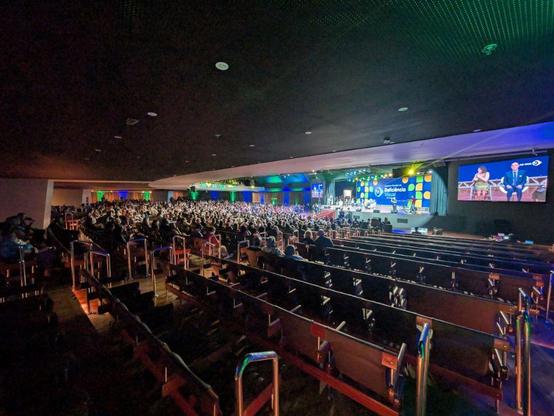 Shot from mid-auditorium, showing the audience in the foreground and a panel on stage. Overhead trusses carry bright spotlights. The left of the stage has a large banner reading “Encontro Mundial da Deficiência Visual.” A side screen shows a person in a blue suit speaking while another panelist sits nearby.