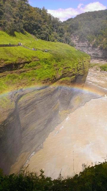 A dramatic gorge cuts through layered sedimentary rock formations, with moss-covered limestone cliffs rising from muddy brown water. A vibrant rainbow arcs across the misty spray where a waterfall cascades from the upper left. Visitors dot the green hillside above, dwarfed by towering trees. The scene is framed by lush vegetation and stratified canyon walls under a partly cloudy sky.