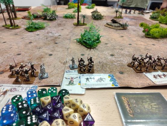 Table de wargame avec figurines. Des figurines et des dés au premier plan, un tapis et des arbres en arrière plan