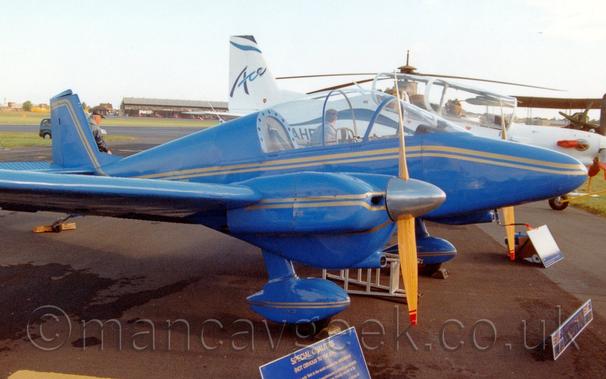 Side view of a twin propellor-engined light aircraft parked on tarmac facing to the right.
The plane is a almost entirely blue, with a double gold stripe running along the body from the nose, as well as along the engine cowlings, just below the wings, and on the spats over the fixed undercarriage, suspended below the engine cowlings.
A large bubble canopy covers the tandem 2-seat cockpit with a person wearing a grey jacket visible through the canopy, standing by the white plane on the far side.
A military green helicopter and a large, twin engined bi-plane are visible in the background on the right, with a black hangar across the far side of the runway on the left, all under hazy grey sky.