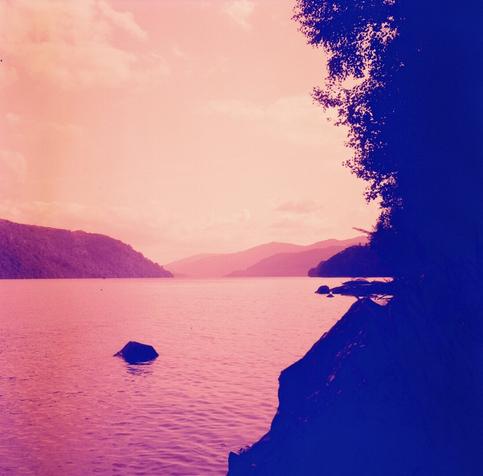 A photo of Loch Lomond shot on a red scale film. The left hand side of the picture shows a red water surrounded by hills in the distance. On the right hand side a tree and some rocks are in a deep blue colour.
The picture is square in shape.