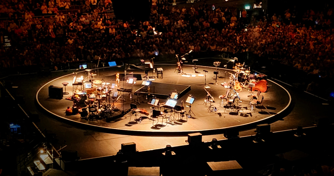 Eine leere Bühne in der Mitte einer Veranstaltungshalle. Die Musiker sind weg. Nur noch ein paar Instrumente und sonstiges Equipment ist zu sehen. Im Hintergrund sind noch einige Zuschauer zu sehen.