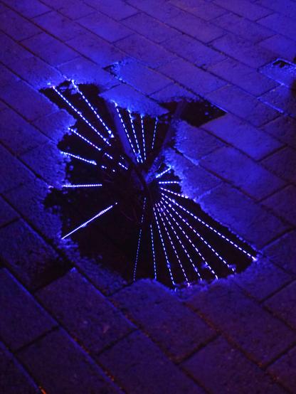 Picture of a water puddle that reflects a middle part of a ferris wheel and its white lights. The pavement is also wet and it reflects blue and pink neon lights.
