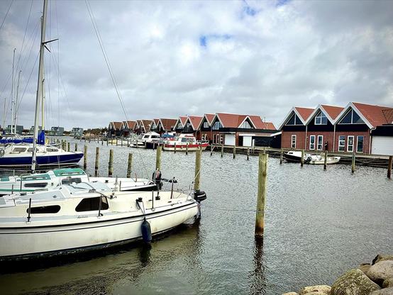Sailboats docked at a marina with calm water, foreground showing a white sailboat closest to the viewer. In the background, a row of red-roofed houses lines the waterfront, under a cloudy sky.