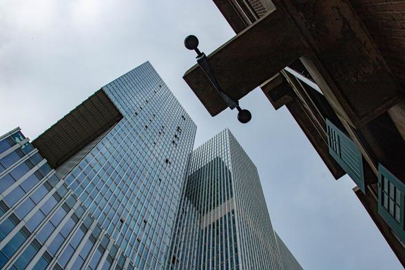 The upper part of two buildings, photographed diagonally from below, with a patch of blue-gray sky between them. Bottom left: a triangle of the same several tall modern buildings, varying in height and depth. Rows of glass windows extending from bottom to top, light bluish-gray because of the reflective sky. The upper right side is dark and shadowed. An old building; a few shutters are visible. A black rectangular section protrudes from the center, with knobs on either side. It fits perfectly between the buildings.