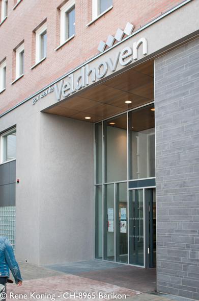 Außeneingang des modernen Gemeindegebäudes in Veldhoven. Das Gebäude zeichnet sich durch eine Mischung aus glatten grauen Wänden, Glaspaneelen, Ziegelmauerwerk im Obergeschoss und einer stilvollen Beschilderung mit der Aufschrift "gemeente Veldhoven" über dem Eingang aus. Der Eingang ist gut beleuchtet, und ein gepflasterter Gehweg führt zu den Glastüren.