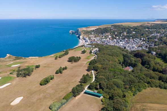 Aerial view of a coastal landscape featuring a golf course, dense greenery, and a seaside town.