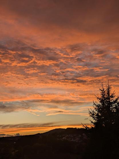 Sonnenuntergang mit lebhaftem Himmel in Orange-, Pink- und Lilatönen. Wolken bilden dynamische Schichten, darunter eine dunkle Landschaft mit Hügeln, Dächern und einem silhouettierten Baum rechts im Bild.