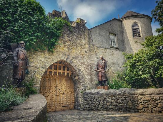 Two large, dark wooden statues of medieval guards stand on either side of a large arched stone gate with a thick, studded wooden door. The statues are positioned in front of a grey stone castle building with a turret on the right. Lush green ivy covers the stone walls of the castle and the surrounding foliage is green and vibrant under a blue, cloudy sky.
