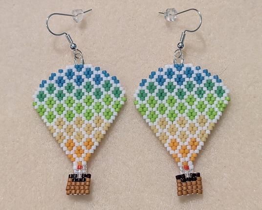 A matching pair of hot air balloon beaded earrings; they ombre from light teal to greens down to yellows and have white between each color. The baskets are standard shades of brown with some iridescent clear beads to represent open air between them and the balloon itself, and there is also a single orange bead on each that's meant to be the flame from their burners.