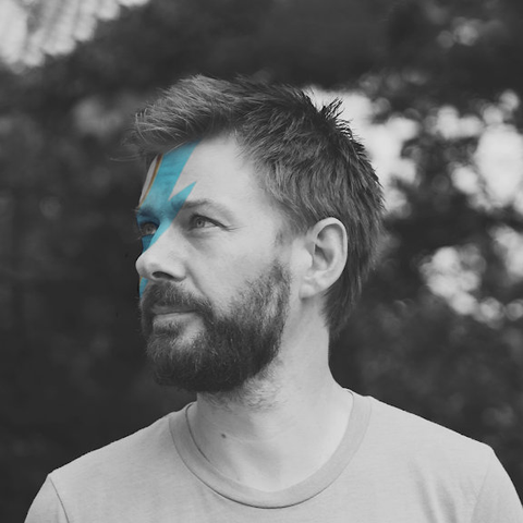 black and white photo of a male with a beard looking to the left standing outside. he has a blue lightning flash painted on his head