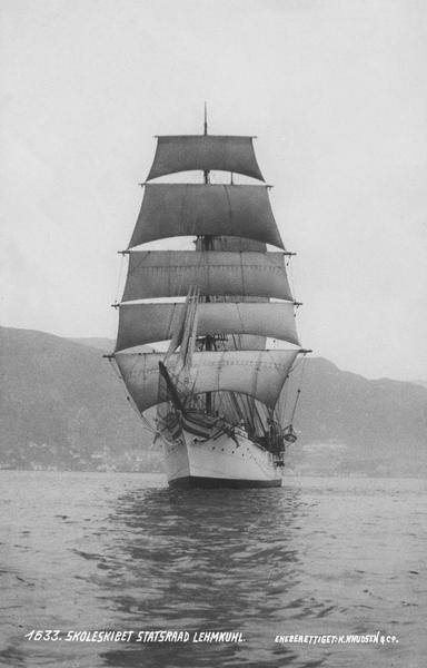 A black and white image of a white three-masted barque with full sails. In the background, mountains.