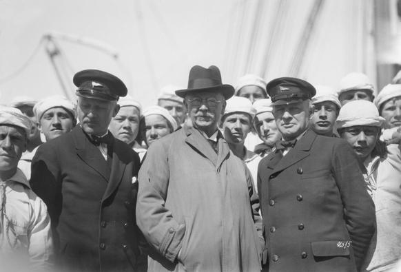 A black and white image of three men in front of ship crew onboard hte Statsraad Lehmkuhl, a chief mate and commander on each side, and centrally a rotund dude with a hat, glasses, and a white moustache – Labour Minister Kristofer Diederich Lehmkuhl.