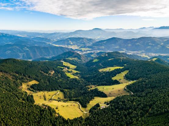Das Bild zeigt eine Landschaftsaufnahme aus der Vogelperspektive. Man sieht sanfte, grüne Wiesen, die von dichten, dunklen Wäldern eingerahmt werden. Kleine, kurvige Straßen schlängeln sich durch die Landschaft. Die Hügel und Berge sind mit einer Mischung aus Waldflächen und offenen Wiesen bedeckt.. Weiter entfernt sieht man mehrere Gebirgsketten, die in bläulichen Tönen erscheinen – ein Effekt, der durch die Atmosphäre entsteht und die Tiefe des Raumes betont. Der obere Teil des Bildes zeigt einen klaren blauen Himmel mit einer Schicht aus weißen Wolken, die teilweise das Sonnenlicht zurückhalten, sodass das Licht sanft und diffus wirkt. Insgesamt vermittelt das Bild eine friedliche, weite und naturbelassene Stimmung – wie eine typische Panoramaansicht in einer alpinen oder voralpinen Region.
