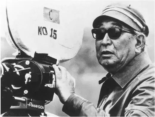 Schwarzweiß Foto von Akira Kurosawa hinter einer Kamera mit der Filmrolle Nr. 15. Kurosawa trägt Mütze und eine Sonnenbrille