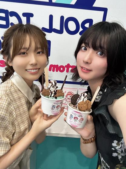 Two women are smiling and posing with cups of rolled ice cream topped with cookies, chocolate drizzle, and biscuits. They are standing in front of a sign with bold blue and pink lettering and anime-style character illustrations. Both women are holding their desserts close to the camera, showcasing the detailed toppings and decorated cups.