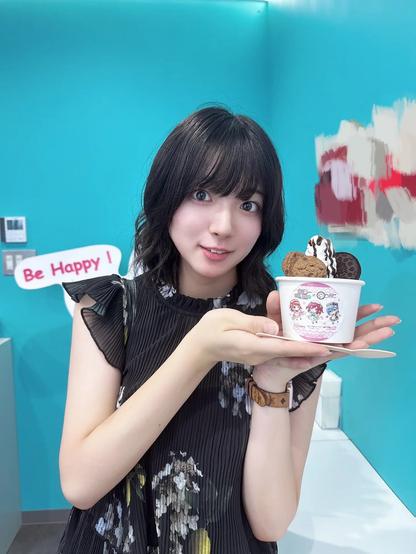 A woman is holding a decorated cup of rolled ice cream topped with cookies, chocolate drizzle, and biscuits. She is standing against a turquoise wall with a sign that says “Be Happy!” in red letters. She is smiling softly at the camera while presenting the dessert with both hands.