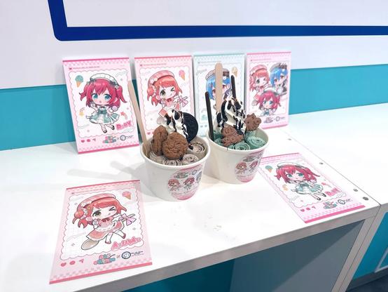 Two cups of rolled ice cream are displayed on a white counter, each topped with cookies, chocolate drizzle, and biscuits. Behind them are colorful cards featuring chibi-style anime characters with bright red and blue hair