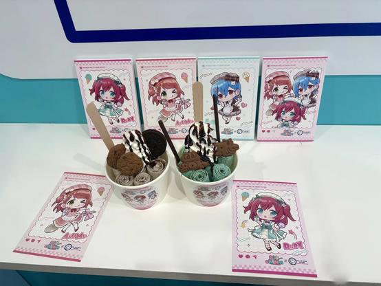 Two cups of rolled ice cream are displayed on a white counter, each topped with cookies, chocolate drizzle, and biscuits. Behind them are colorful cards featuring chibi-style anime characters with bright red and blue hair