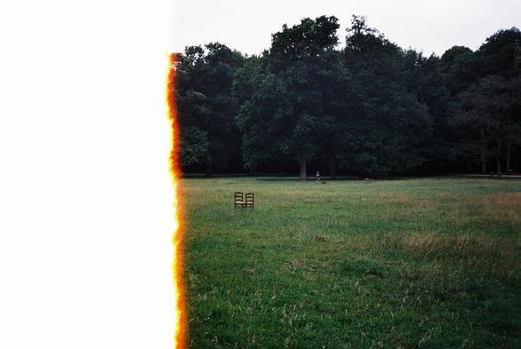 Grassy field with scattered chairs, bordered by tall trees; partial image burn on the left adds dramatic effect.