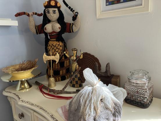 The top of a chest of drawers in the corner of a room is filled with a very large crocheted Minoan Snake Goddess statue, a small Snake Goddess figurine, a gold labrys, a pair of brown sacred horns, a bowl of dried palm leaves on a gold offering stand, a rustic sistrum, a pressed glass jar, a finger labyrinth, and a white plastic garbage bag containing a large lump of raw clay.