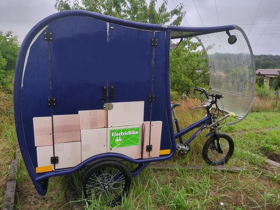 Ein CargoTrike, mit blauer Folierung, das mal von einem Paketdienst genutzt wurde, steht auf einer Rasenfläche. 
Da es stark geregnet hatte, ist das Fahrzeug nass. 
Über dem Sitzbereich verläuft das Dach, an dem eine Frontscheibe montiert ist.