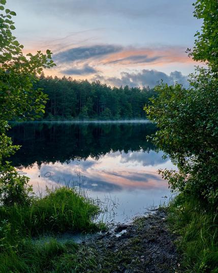 A tranquil scene of a lake surrounded by lush greenery and trees, reflecting the colorful sky at dusk. Soft clouds and hints of pink and blue characterize the evening sky, creating a serene atmosphere.