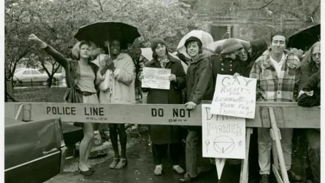 Foto en blanco y negro de varias personas tras una vaya policial en la que pone "POLICE LINE". Las personas visten al estilo de los años 70 en Estados Unidos. Aunque está lloviendo (a juzgar por varios paraguas que salen), su carácter es alegre, y sostienen varios carteles con consignas (que no consigo leer bien), pero que van a favor de los derechos para el colectivo gay. En este grupo de gente ( en el que me parece contar 12 cabezas), a la izquierda del todo, se encuentran Sylvia Rivera y Marsha P. Jhonson Sylvia aparece a la izquierda, con un vestido corto de color claro ceñido por un cinturón negro a la cintura, y lleva una chaqueta que parece cuero así como un bolso (ambos de colores oscuros). Aparece levantando el puño derecho en señal de protesta. Marsha aparece a la derecha, con un chaquetón que parece de "pelo", con un bolso blanco. Sujeta un paraguas con el que resguarda a ambas. Tiene una flor en el pelo. Esta es la foto en la que me baso para hacer el dibujo.