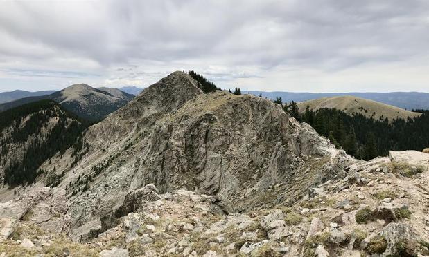 A view from a mountain peak, looking across to another peak of similar height. We're above the treeline, so the ground is covered with jagged rocks and no trees, though there's a little bit of lichen and low growth between the rocks. In the distance you can see a few other peaks, slightly lower, with grassy bald spots at the top and trees lower. The sky is overcast.