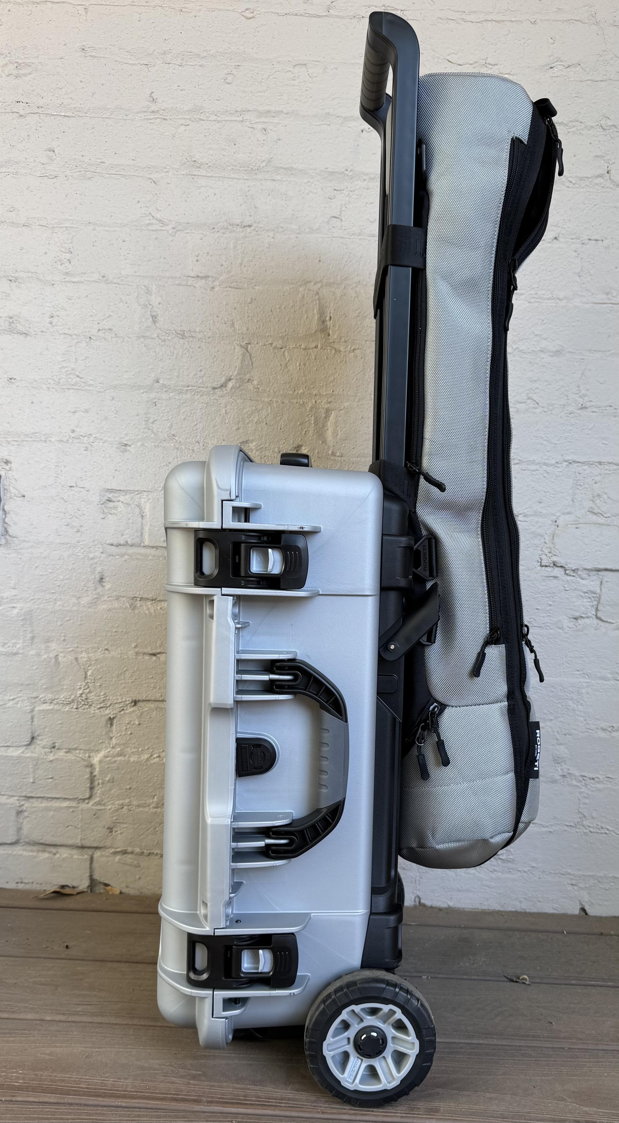 A grey rolling carryon suitcase with large wheels and a tripod bag slipped over the carrying handle