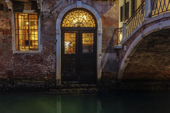 An old brick house by a canal in Venice (Italy) illuminated at night from inside. A golden light comes out the iron work top of a beautiful  door and from a slightly open window revealing a beautiful bookshelve. On the right a stone bridge illuminated from above by the street lights. The canal is of a dark green color. The whole atmosphere is peaceful and mysterious.