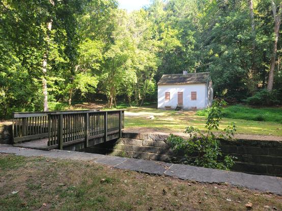 The lock and its stone facing crosses the frame from left to right. A wooden footbridge crosses the lock at the left. An open grassy area, the lockhouse and trees in the background.