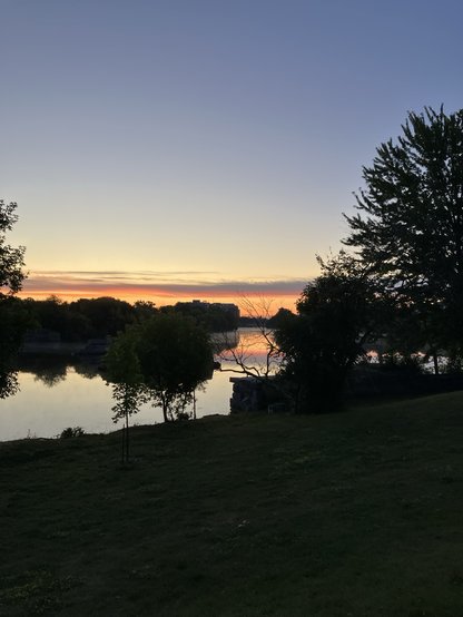 Orange glow at the horizon reflecting on the underside of clouds and on the water. Silhouette of trees creating negative space with the ombré grey sky