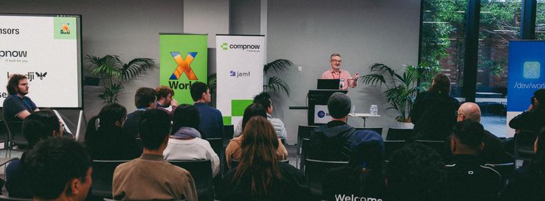 A man standing at a podium, addressing a crowd at a conference. Banners for X World and /dev/world are around him.