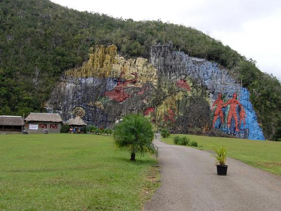A hillside of vegetation has been stripped down to solid rock in one large section where a massive mural has been painted. The mural shows two adults and a child walking, dinosaurs and colors of blue, yellow and black fill the space. In front of the mural is a vast lawn and a few very small buildings. Vinales, Cuba