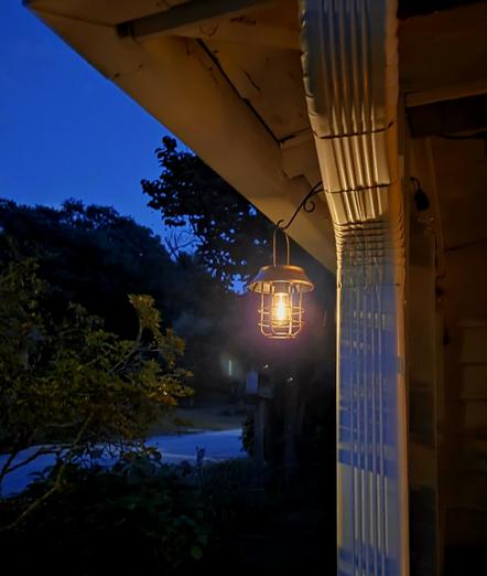 The photo shows a warmly glowing porch light hanging from the corner of a house at dusk. The light fixture has a rustic, old-fashioned design with a wire cage surrounding the bulb and a metal cap on top, giving it a vintage lantern look. It’s attached to the house by a curled metal bracket, hanging just beneath the eaves beside a white downspout. The background fades into deep blue evening sky and the shadowy outlines of trees and shrubs, with a faint glimpse of a quiet road. The scene feels calm, cozy, and a little nostalgic like the soft welcome of a mountain evening in Appalachia settling in.