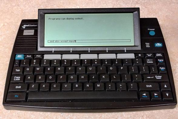 Cidco MailStation (black keyboard with built-in grayscale LCD screen).  The top of the screen reads "Programs can display output...".  A rectangular field near the bottom of the screen reads "...and also accept input", with a vertical bar cursor.