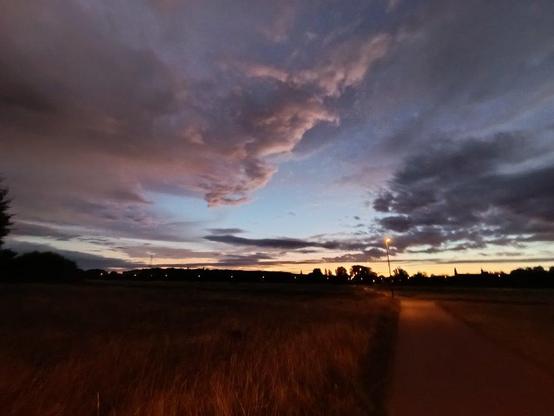 Weitwinkel, Wolkenhimmel von unten angestrahlt 
 Blau Grau rosa
Darunter Wiesen und Radweg.
Baumsilhouetten an Horizont
