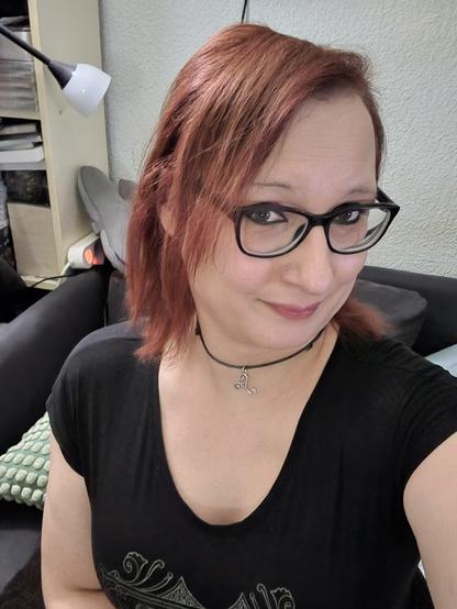 Das Bild zeigt eine Frau mit roten Haaren, die eine schwarze Brille trägt und ein schwarzes T-Shirt mit einem dekorativen Muster trägt. Sie sitzt auf einem schwarzen Sofa und trägt eine schwarze Halskette mit einem silbernen Anhänger. Im Hintergrund ist ein weißer Lampenschirm und ein Bücherregal zu sehen, das mit Büchern und anderen Gegenständen gefüllt ist. Die Frau lächelt leicht in die Kamera.