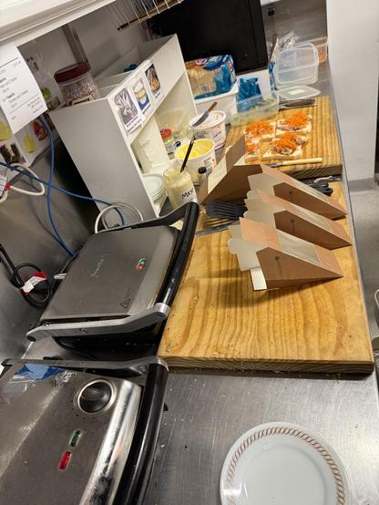 A kitchen bench with sandwich toastie makers and cutting boards, and sandwiches about to be packaged up.