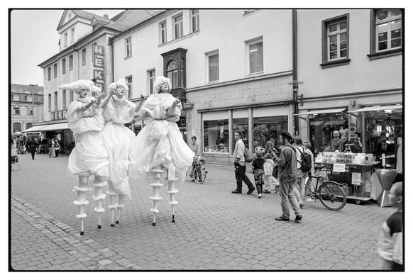 Black and white photograph in landscape orientation.
Three actors on stilts  with white costumes are standing in a street with some people looking interested while others are minding their own business.