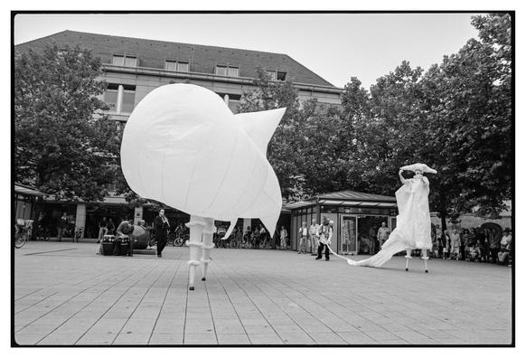 Black and white photograph in landscape orientation.
Another actor has entered the scene and is inflating the costumes of two of the original actors. The first one is  now wear a balloon like costume with 2-3 meters diameter. The second one is being filled up.