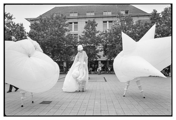 Black and white photograph in landscape orientation.
The two actors with their inflated costumes are proudly parading on the square. The third actor is sadly standing between them.