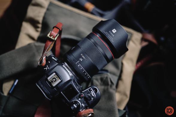 A Canon camera with a 24mm lens rests on a green canvas backpack with leather straps.