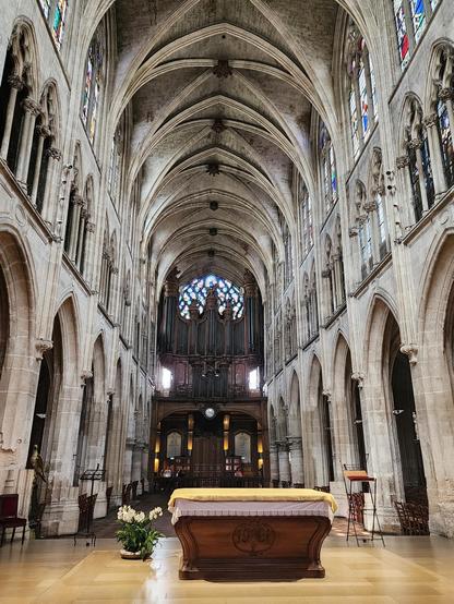 Looking down the center of the cathedral towards the large organ high in the far back, with the altar in the foreground. The ceiling is ribbed and vaulted, and the walls on either side are composed of large stone columns on the bottom level and two levels of stained glass windows.