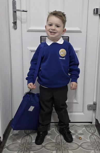 A young boy stands in front of a white door, smiling. He is wearing a blue school sweater with a school logo, a white collared shirt, and dark trousers. He holds a blue bag over his shoulder and has short light brown hair.
