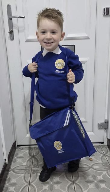 A young boy is smiling while holding a blue school bag over his shoulder. He is wearing a blue school sweater with a logo, standing in front of a white door, with patterned flooring visible.